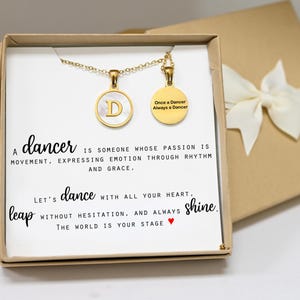 May include: A gold-colored necklace with two round pendants, one with the letter "D" and the other with the text "Once a Dancer Always a Dancer." The necklace is presented in a gift box with a white bow. The box contains a card with text about dance.