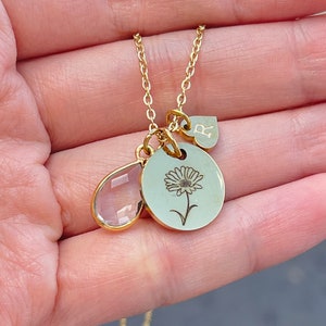 May include: A gold-colored necklace with three charms. One charm is a teardrop-shaped, clear crystal. Another is a round charm with a daisy engraving. The third charm is a heart-shaped charm with the letter "R".