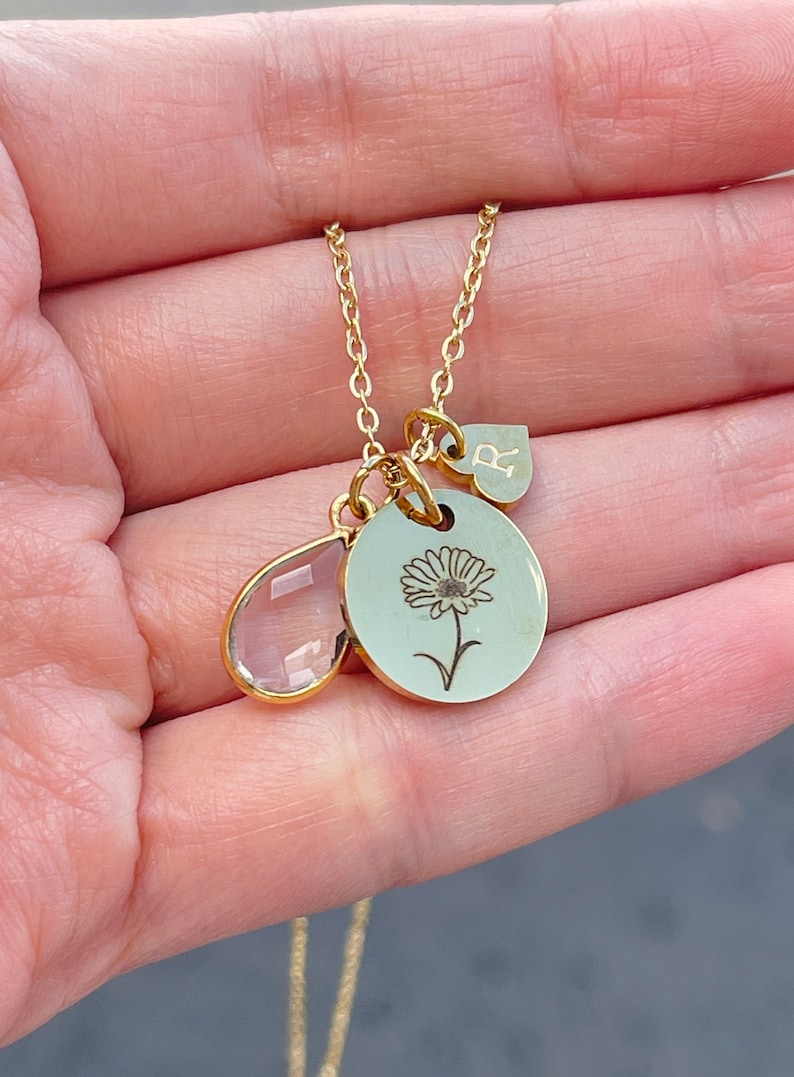 May include: A gold-colored necklace with three charms. One charm is a teardrop-shaped, clear crystal. Another is a round charm with a daisy engraving. The third charm is a heart-shaped charm with the letter "R".