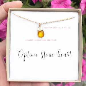 Op de afbeelding: Een gouden ketting met een traanvormige edelsteen hanger en een klein hartje met de letter 'K' erop. De ketting zit in een witte doos met de tekst '(CUSTOM INITIAL A TO Z)' en '(CUSTOM BIRTHSTONE JAN-DEC)' erop. De tekst 'Option Stone heart' staat ook op de doos.