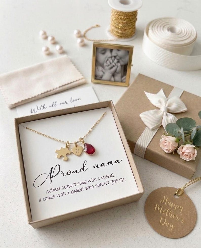 May include: A gold necklace with puzzle piece, heart, and teardrop charms, presented in a gift box with a card that reads "Proud mama." The image includes a small photo frame, a gift box with a bow, and a tag that says "Happy Mother's Day."