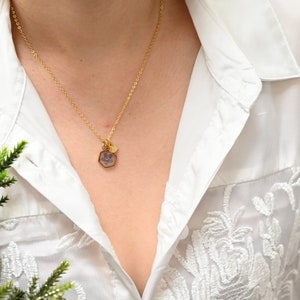 K&ouml;nnte beinhalten: Eine goldene Kettenkette mit einem kleinen sechseckigen Anh&auml;nger mit einer gepressten lila Blume. Die Kette wird von einer Person getragen, die eine wei&szlig;e Bluse mit zarten Stickereien tr&auml;gt.