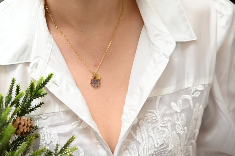 K&ouml;nnte beinhalten: Eine goldene Kettenkette mit einem kleinen sechseckigen Anh&auml;nger mit einer gepressten lila Blume. Die Kette wird von einer Person getragen, die eine wei&szlig;e Bluse mit zarten Stickereien tr&auml;gt.