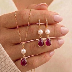 May include: A gold necklace and earrings set featuring pearls and teardrop-shaped ruby gemstones. The necklace has a single pearl and ruby pendant. The earrings have a pearl and ruby drop design, with small clear stones on the earring hooks.