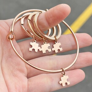 May include: Rose gold-colored bracelets with puzzle piece charms. The bracelets are a mix of adjustable bangles and simple bands. The charms are small puzzle pieces, and the bracelets are held in a hand.