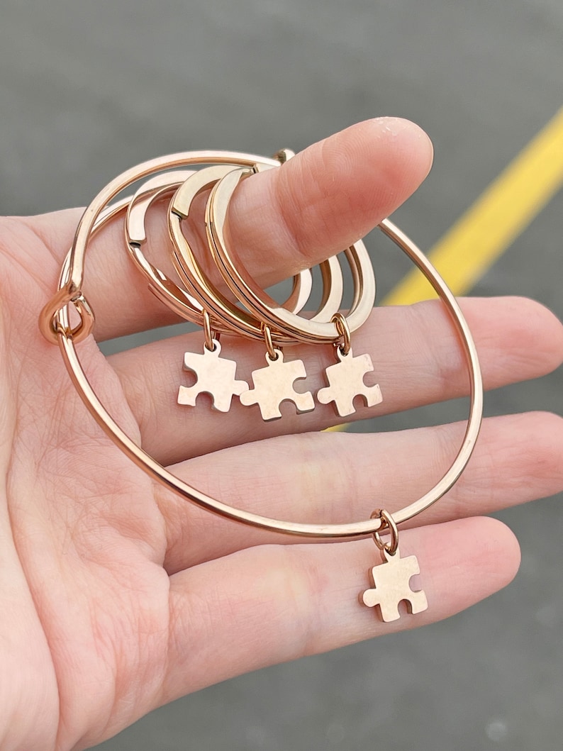 May include: Rose gold-colored bracelets with puzzle piece charms. The bracelets are a mix of adjustable bangles and simple bands. The charms are small puzzle pieces, and the bracelets are held in a hand.