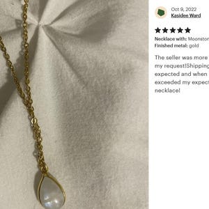 May include: A gold chain necklace with a teardrop-shaped moonstone pendant. The necklace is on a white fabric background.