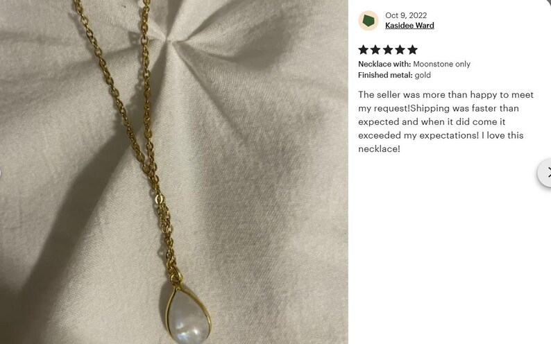 May include: A gold chain necklace with a teardrop-shaped moonstone pendant. The necklace is on a white fabric background.