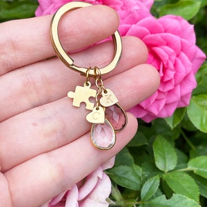 May include: A gold-colored keychain with a puzzle piece charm, two heart-shaped charms with the letters "C" and "K", and two teardrop-shaped crystal charms. The keychain is held in front of pink roses.