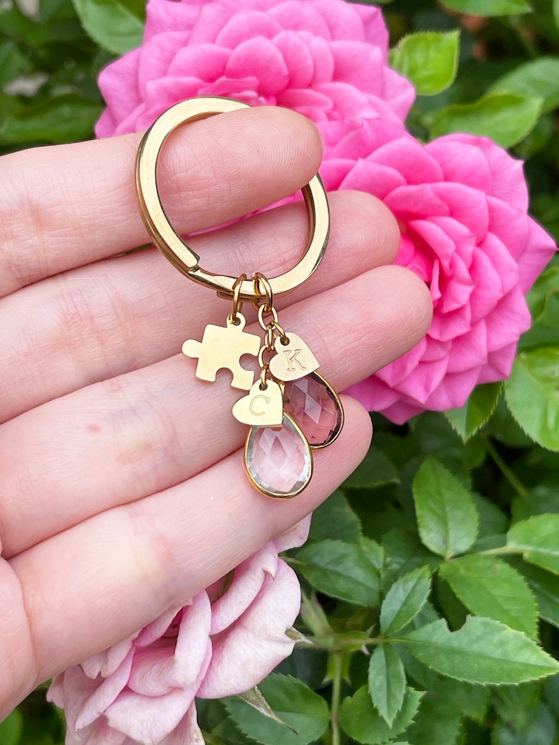 May include: A gold-colored keychain with a puzzle piece charm, two heart-shaped charms with the letters "C" and "K", and two teardrop-shaped crystal charms. The keychain is held in front of pink roses.
