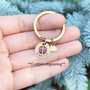Op de afbeelding: Gouden sleutelhanger met een ronde hanger met een rood kruis en de tekst "JOHN 3:16". Een kleine hartvormige hanger is aan de ring bevestigd.