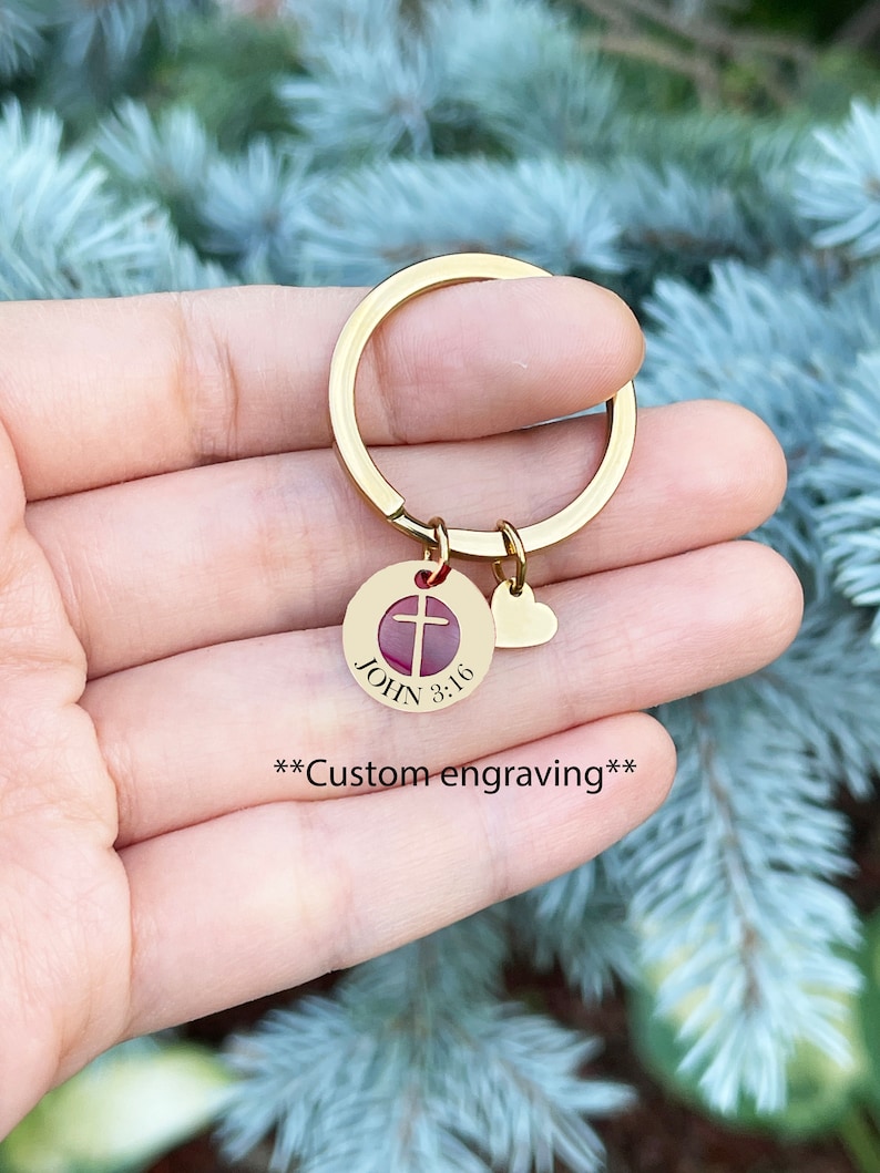 Op de afbeelding: Gouden sleutelhanger met een ronde hanger met een rood kruis en de tekst "JOHN 3:16". Een kleine hartvormige hanger is aan de ring bevestigd.