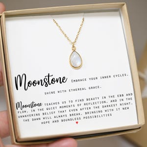 May include: A gold necklace with a teardrop-shaped moonstone pendant in a white box. The box has a message about moonstone: "Moonstone embraces your inner cycles, shine with ethereal grace. Moonstone teaches us to find beauty in the ebb and flow, in the quiet moments of reflection, and in the unwavering belief that even after the darkest night, the dawn will always break, bringing with it new hope and boundless possibilities."