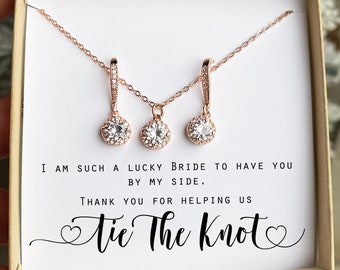May include: A white box with a rose gold necklace and earrings set. The box has a message that reads "I am such a lucky bride to have you by my side. Thank you for helping us tie the knot."