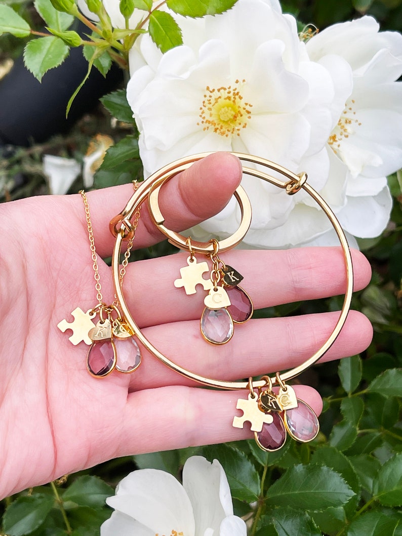 May include: A gold-colored jewelry set featuring a necklace, a keychain, and a bangle bracelet. Each piece has puzzle piece charms and teardrop-shaped gemstones in shades of pink and clear. The jewelry is held in front of white flowers.