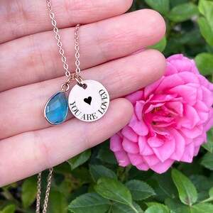 May include: A rose gold necklace with two pendants. One pendant is a teardrop-shaped blue gemstone, and the other is a round charm with the words "YOU ARE LOVED" and a black heart. The necklace is displayed against a backdrop of pink roses and green foliage.