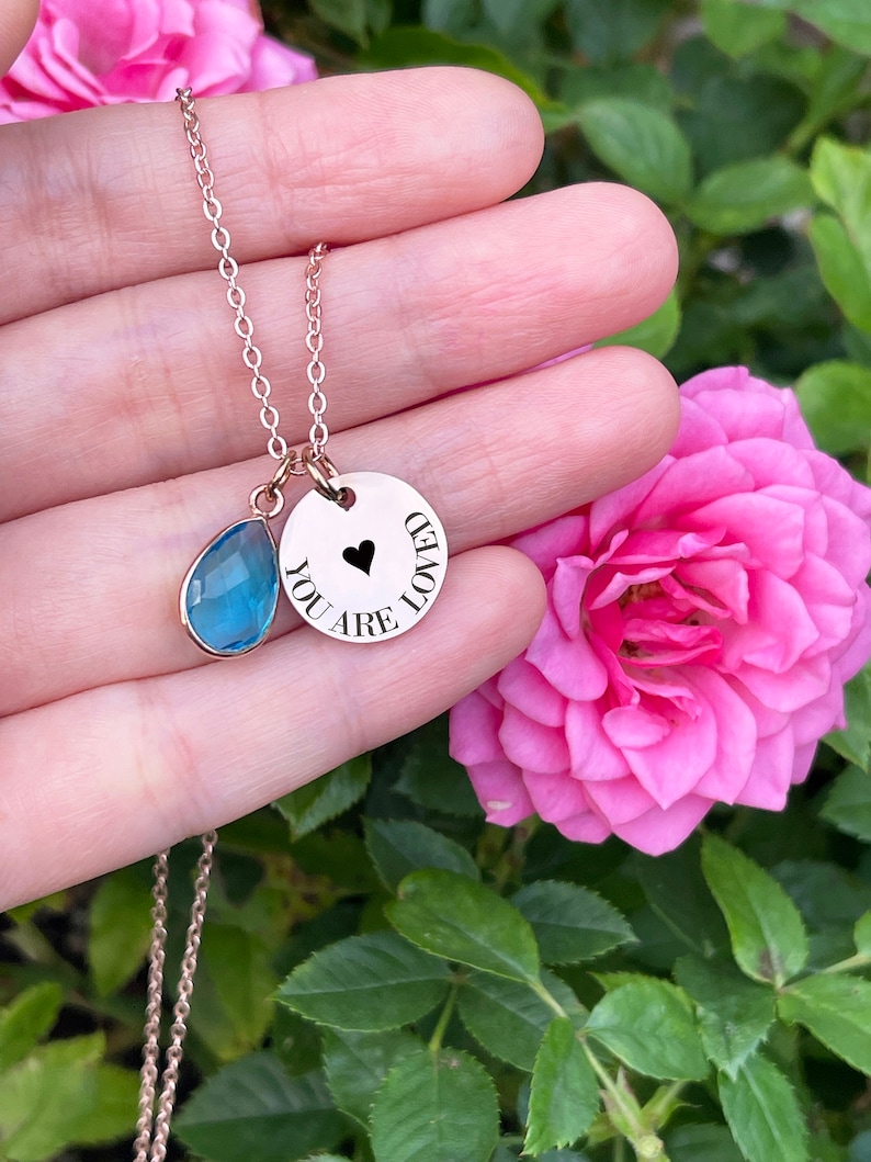May include: A rose gold necklace with two pendants. One pendant is a teardrop-shaped blue gemstone, and the other is a round charm with the words "YOU ARE LOVED" and a black heart. The necklace is displayed against a backdrop of pink roses and green foliage.