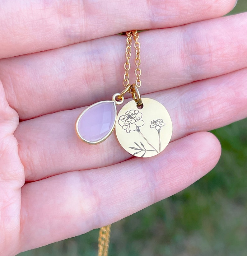 May include: A gold-colored necklace with two pendants. One pendant is a teardrop-shaped, light pink stone. The other is a round, gold-colored pendant with a floral design. The necklace is on a gold chain.