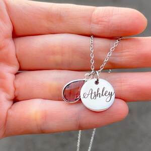 May include: A silver necklace with a teardrop-shaped purple gemstone and a round pendant engraved with the name "Ashley".