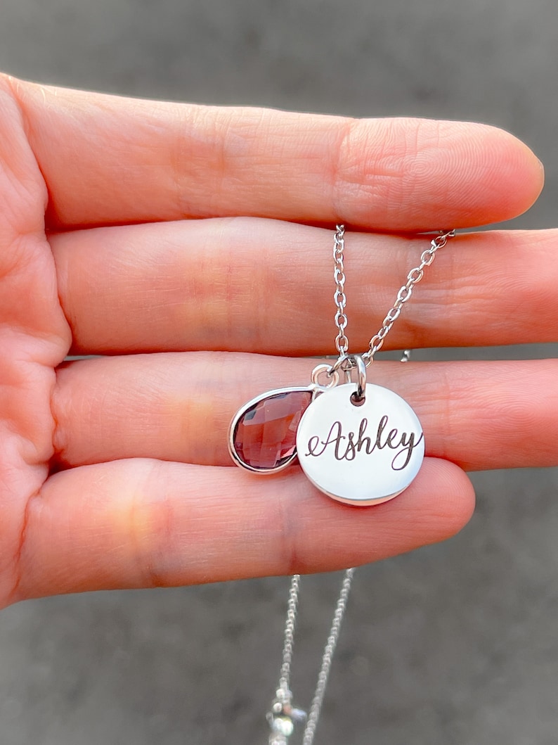 May include: A silver necklace with a teardrop-shaped purple gemstone and a round pendant engraved with the name "Ashley".
