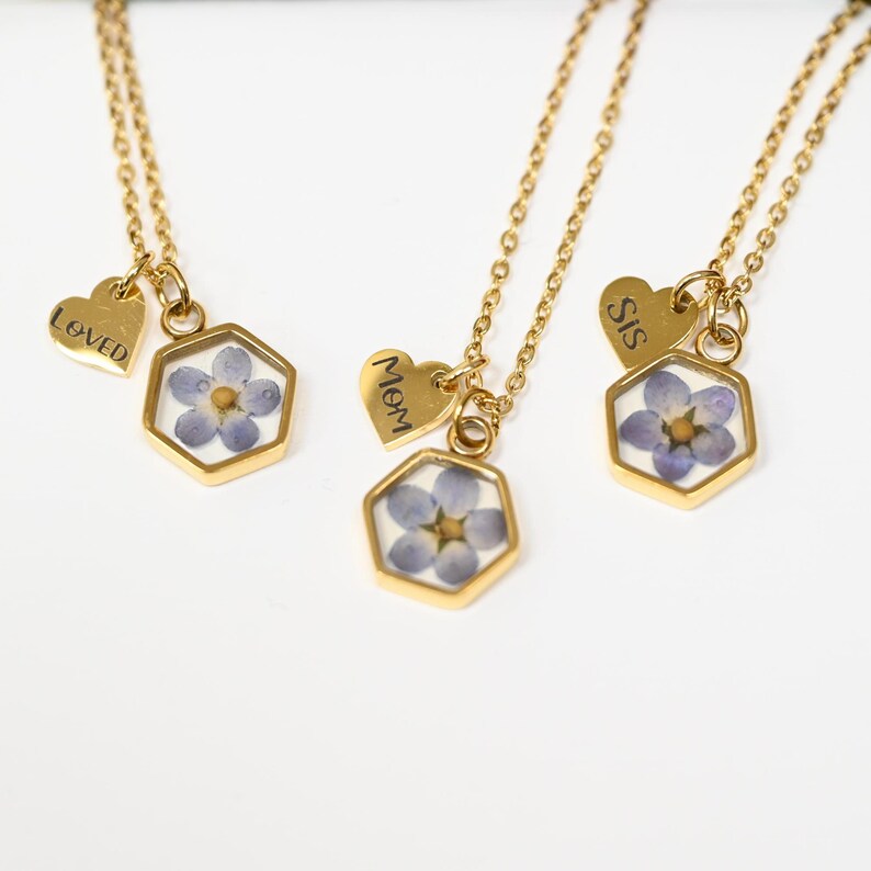 May include: Gold necklaces featuring hexagonal pendants with blue flowers. Each necklace has a small heart charm with the words "Loved", "Mom", or "Sis". The chains are delicate and the pendants are gold-framed.