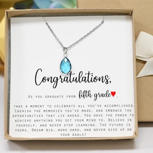 May include: A silver necklace with a teardrop-shaped, light blue pendant, presented in a gift box. The card inside reads "Congratulations" and "fifth grade" with a heart symbol, along with an inspirational message.