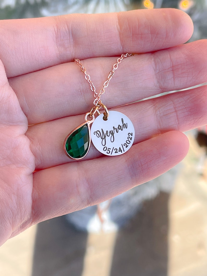 May include: A gold necklace with a green teardrop-shaped gemstone and a round pendant with the name "Veyrah" and the date "05/24/2022" engraved on it.