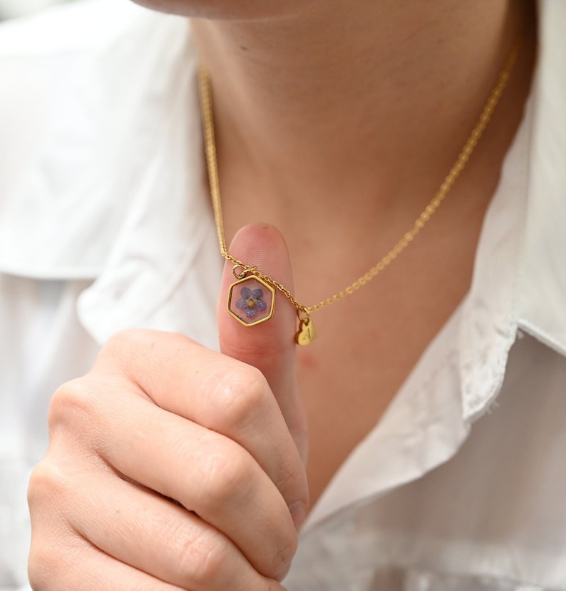 May include: A gold necklace with a hexagonal pendant containing a small purple flower. The chain is fine, and there's a small heart-shaped charm. The background is a white shirt.