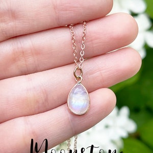 May include: A delicate rose gold chain necklace with a teardrop-shaped moonstone pendant. The stone has a milky white color with iridescent flashes of blue and green.