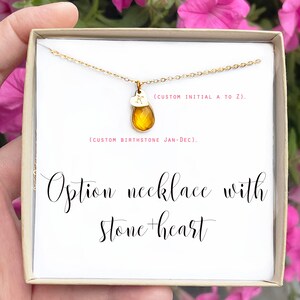 May include: A gold-colored necklace with a teardrop-shaped amber stone and a small heart pendant with the letter "K". The necklace is presented in a gift box. Text on the box reads "Option necklace with stone + heart".