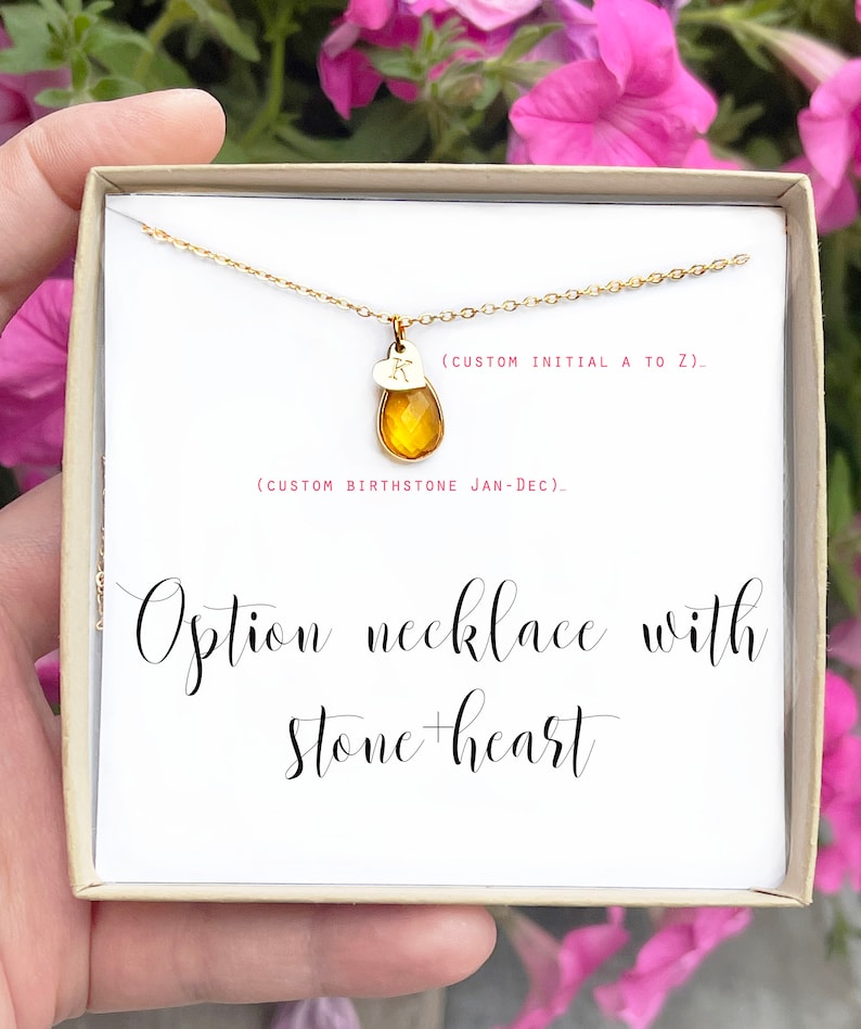 May include: A gold-colored necklace with a teardrop-shaped amber stone and a small heart pendant with the letter "K". The necklace is presented in a gift box. Text on the box reads "Option necklace with stone + heart".