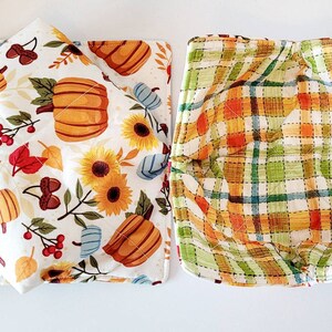 May include: Two fabric bowl cozies. One features a white background with pumpkins, sunflowers, and fall foliage. The other has a plaid pattern in orange, green, and yellow. Both are quilted.