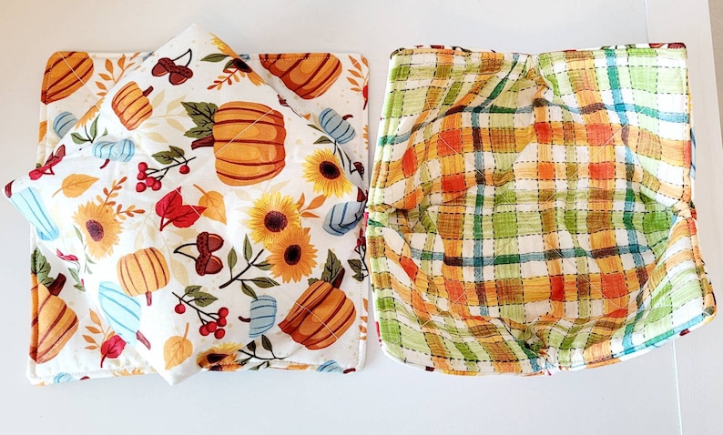 May include: Two fabric bowl cozies. One features a white background with pumpkins, sunflowers, and fall foliage. The other has a plaid pattern in orange, green, and yellow. Both are quilted.