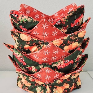May include: A stack of five fabric bowl cozies. The cozies have a red and green pattern with pumpkins, snowflakes, and red trucks. The interior is a solid red with white snowflakes. The cozies are stacked on a white surface.