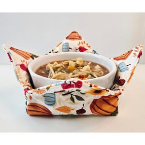May include: A fabric bowl cozy featuring a cream background with a colorful print of orange pumpkins, blue pumpkins, red and yellow autumn leaves, and brown acorns, holding a white ceramic bowl filled with a vegetable noodle soup.