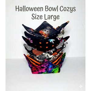 May include: A stack of large fabric bowl cozies featuring various Halloween-themed patterns including skulls, orange polka dots, and colorful neon prints. The text "Halloween Bowl Cozys Size Large" is printed in dark gray above the fabric items.