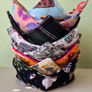 May include: A stack of six colorful fabric bowl cozies. The cozies are made of different fabrics with various patterns, including butterflies, plaid, and sunflowers.