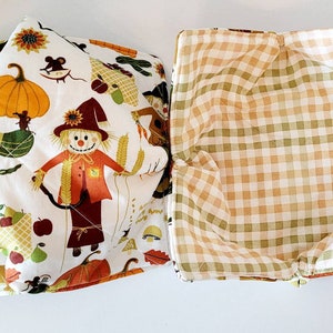 May include: Two fabric bowl cozies. One features a white background with a fall harvest print of pumpkins, scarecrows, and mice. The other has a tan and green plaid pattern. These are designed to keep bowls warm.