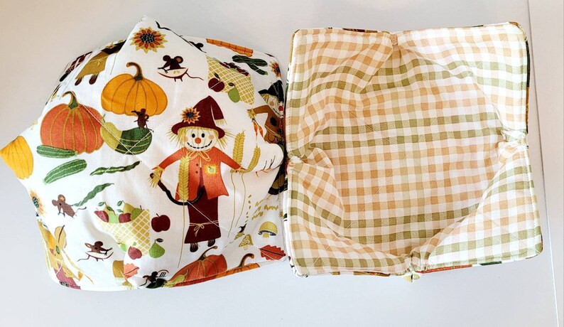 May include: Two fabric bowl cozies. One features a white background with a fall harvest print of pumpkins, scarecrows, and mice. The other has a tan and green plaid pattern. These are designed to keep bowls warm.