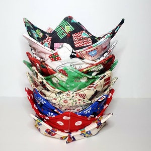 May include: A stack of eight fabric bowl cozies in various Christmas patterns, including red, green, blue, and white. The cozies are folded in a bowl shape and stacked on top of each other.