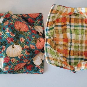 May include: Two quilted bowl cozies. One is teal with orange and white pumpkins, flowers, and leaves. The other is a plaid pattern in orange, green, and yellow. These are designed to protect hands from hot bowls.