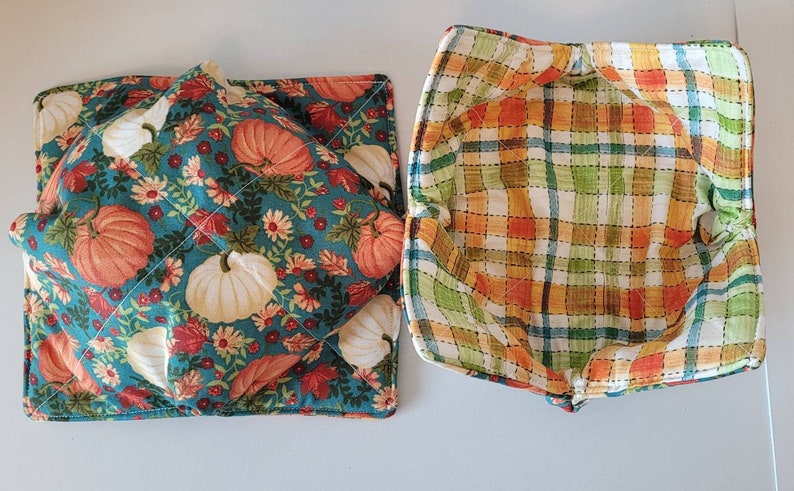May include: Two quilted bowl cozies. One is teal with orange and white pumpkins, flowers, and leaves. The other is a plaid pattern in orange, green, and yellow. These are designed to protect hands from hot bowls.