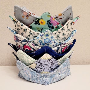 May include: A stack of six fabric bowl cozies in various floral patterns, including blue, green, and white. The cozies are folded into a crown shape.