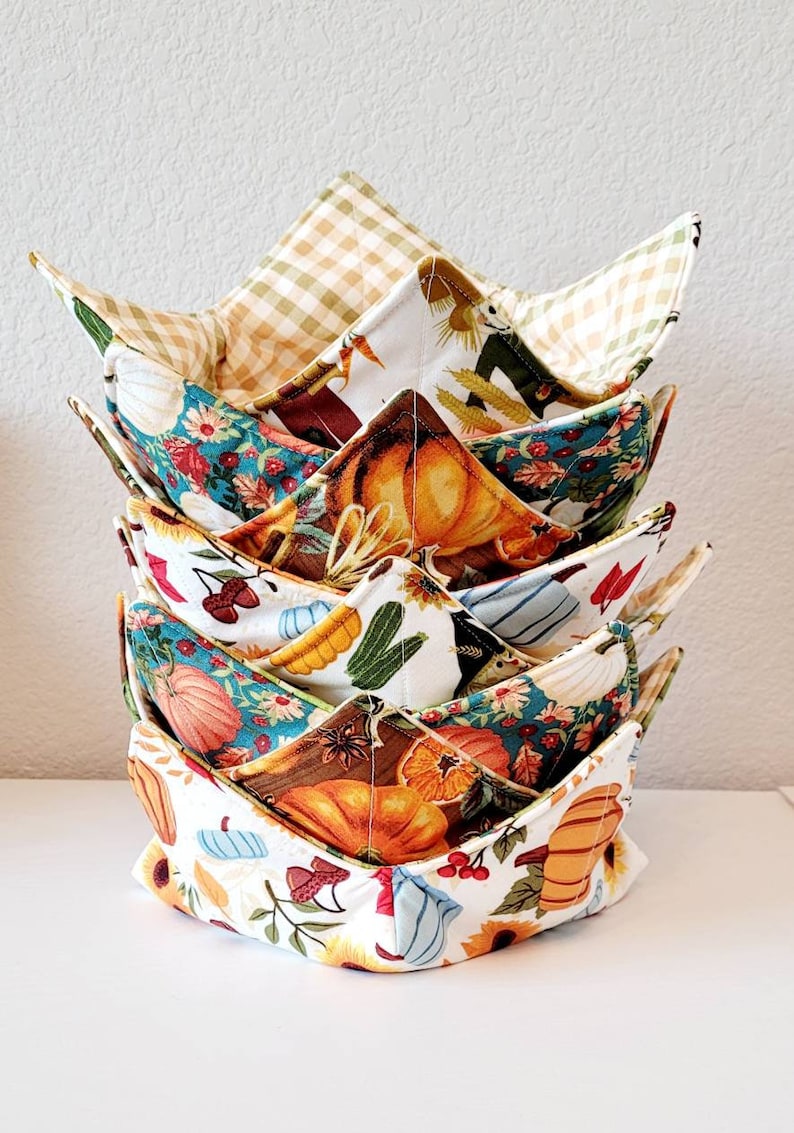 May include: A stack of six fabric bowl cozies with various fall-themed patterns. Designs include pumpkins, sunflowers, and plaid. The cozies are in shades of orange, teal, brown, and cream. They are designed to be used in the microwave.