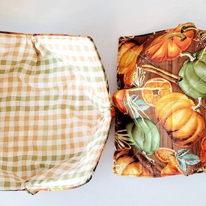 May include: Two quilted bowl cozies. One features a brown, orange, and green pumpkin print on a wood-grain background. The other has a tan, green, and white gingham pattern. Both have a square shape.
