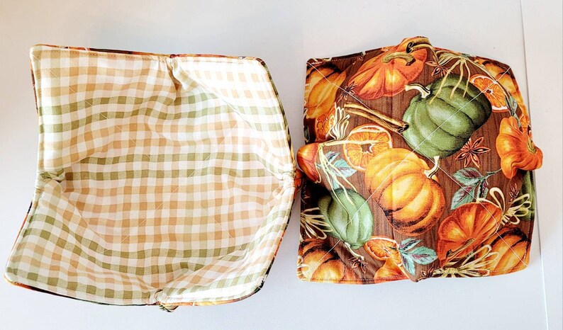 May include: Two quilted bowl cozies. One features a brown, orange, and green pumpkin print on a wood-grain background. The other has a tan, green, and white gingham pattern. Both have a square shape.
