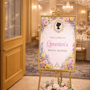 May include: A gold easel with a sign that reads "Welcome to Genevieve's Bridal Shower." The sign is decorated with floral arrangements and a silhouette. A floral arrangement of pink, white, and yellow flowers is at the base of the easel.