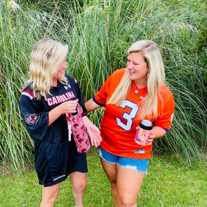以下が含まれることがあります： 2人の女性が大学フットボールのジャージを着ています。一人は黒地に「CAROLINA」と書いてあり、もう一人はオレンジ地に「3」と書いてあります。カラフルなデザインのドリンククーラーを持っています。