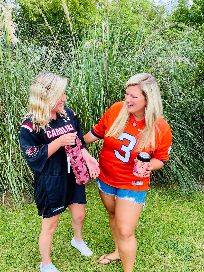 以下が含まれることがあります： 2人の女性が大学フットボールのジャージを着ています。一人は黒地に「CAROLINA」と書いてあり、もう一人はオレンジ地に「3」と書いてあります。カラフルなデザインのドリンククーラーを持っています。