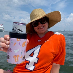 以下が含まれることがあります： 白い数字の4が書かれたオレンジ色のジャージと麦わら帽子をかぶった女性が、ホワイトクローブの缶が入ったピンクと白の缶クーラーを持っています。女性は笑顔でカメラを見ています。